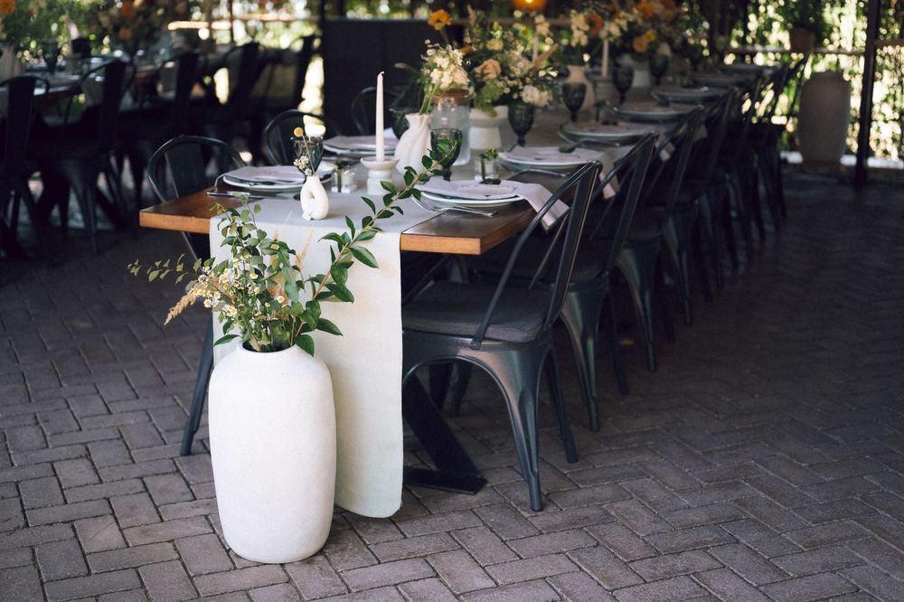 Elegant outdoor dining table with metal chairs, white vases, greenery, and candles on a brick patio.