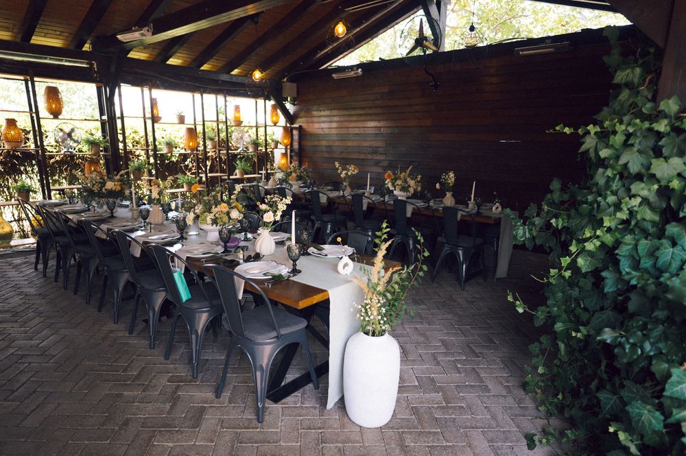 Elegant outdoor restaurant setup with long dining tables, lush greenery, and stylish decor under a covered patio.