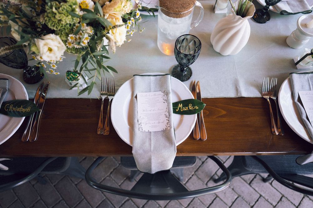 Elegant wedding table setting with white plates, copper cutlery, floral centerpiece, and green place cards on wooden runner.