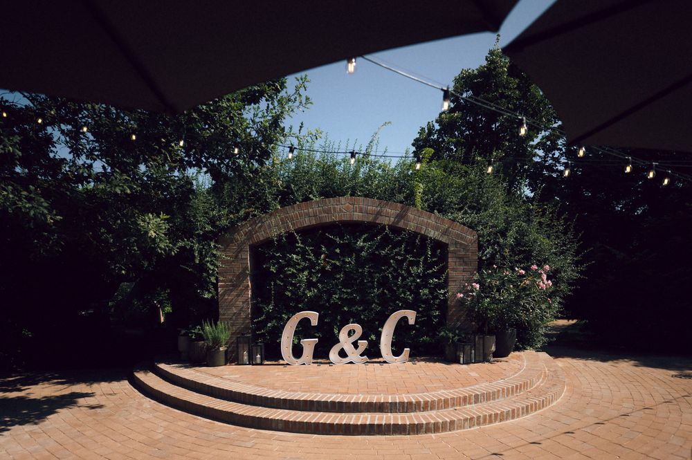 Outdoor wedding venue with large G&C monogram letters on brick steps surrounded by lush greenery and string lights.