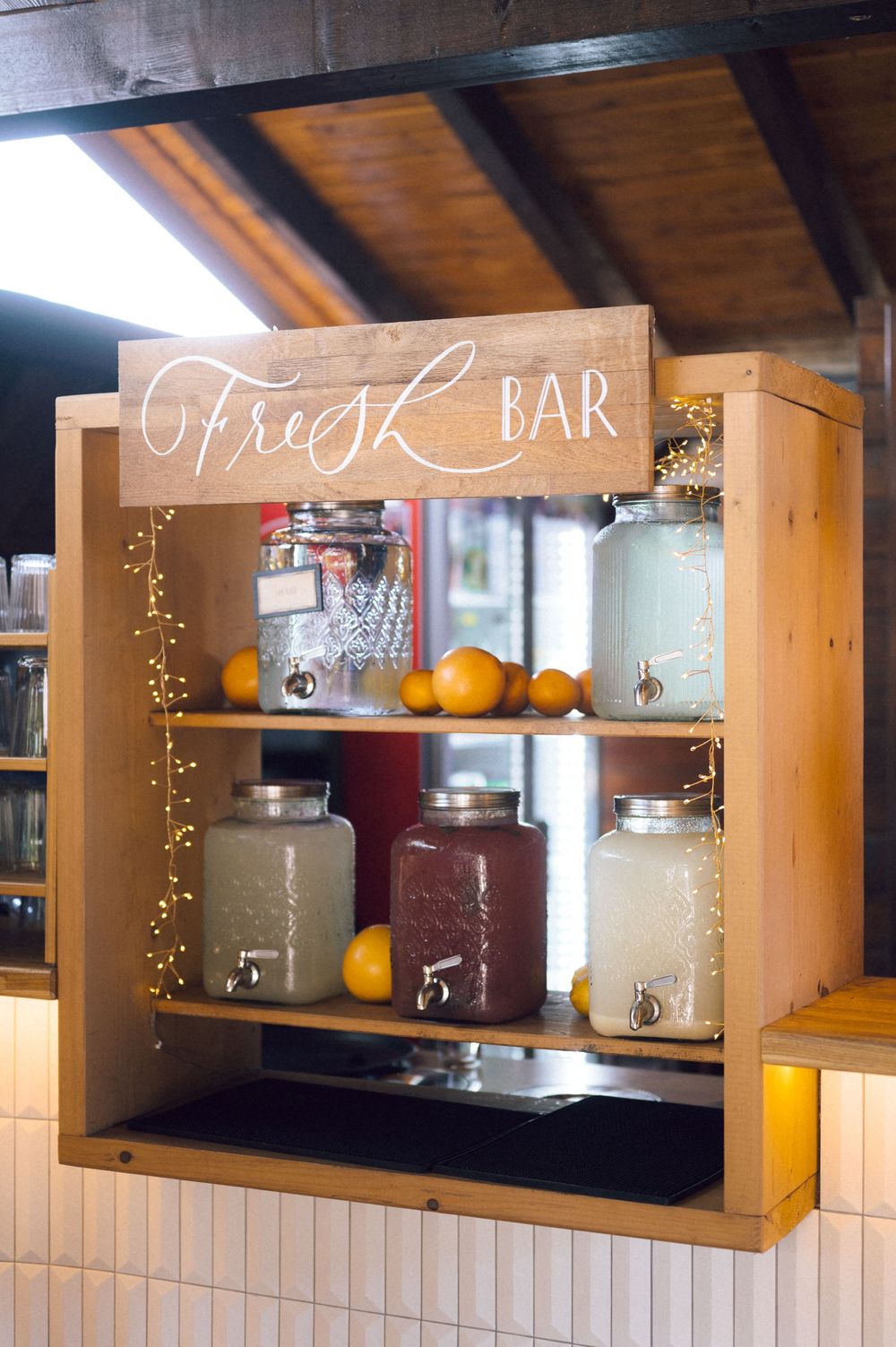 Rustic wooden Fresh Bar display with drink dispensers, fresh oranges, and fairy lights on two shelves.