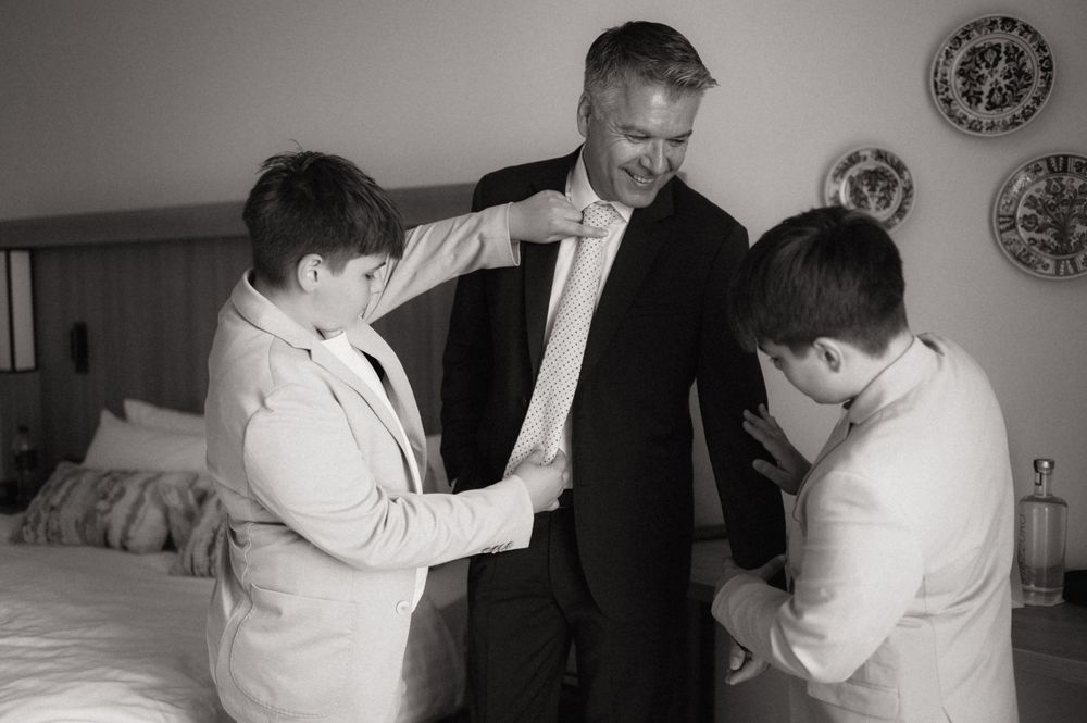 Two boys help a man in a suit adjust his tie and jacket in a bedroom, preparing for a formal wedding celebration.