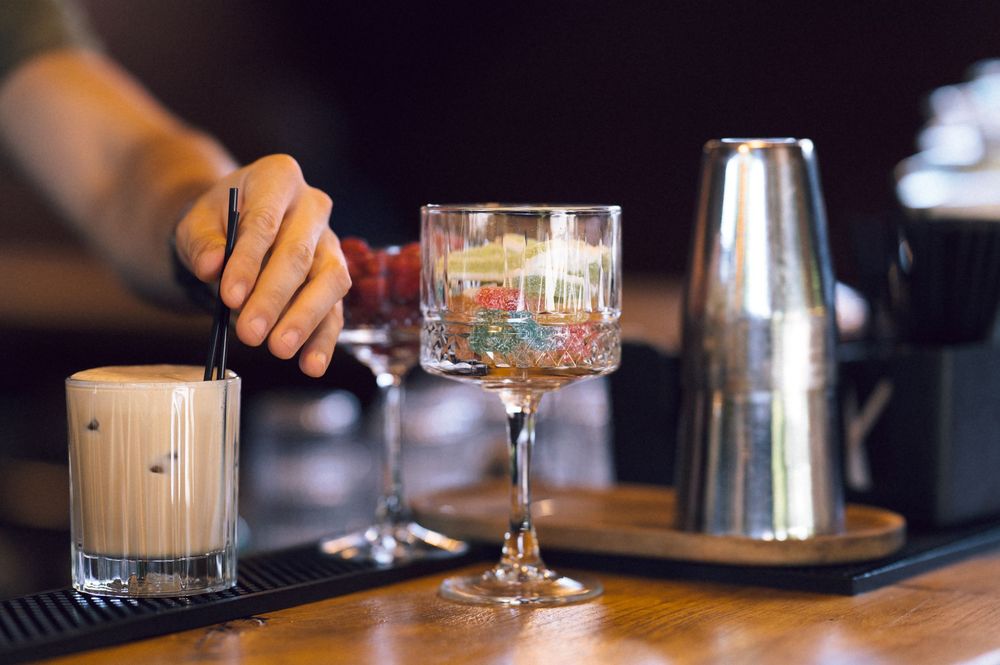 Bartender stirring a creamy cocktail beside an ornate glass and metal shaker on a wooden bar.