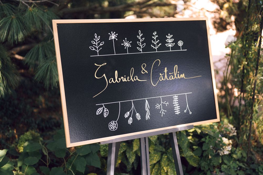 Chalkboard wedding sign on easel reading Gabriela & Catalin with floral doodles, displayed outdoors among green trees.
