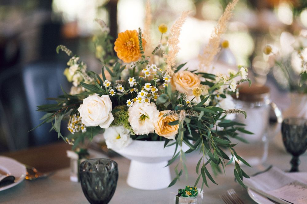 Elegant wedding centerpiece with white and peach roses, yellow marigolds, and greenery in a white pedestal vase on a table.