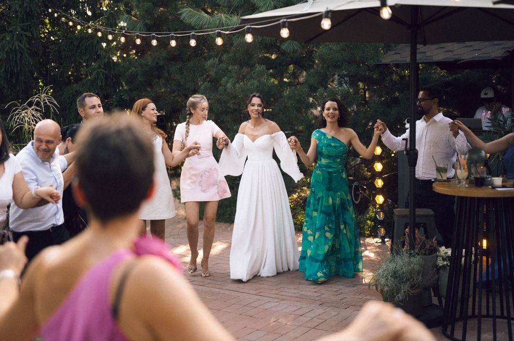 Bride in white gown dancing with guests at outdoor wedding reception under string lights.