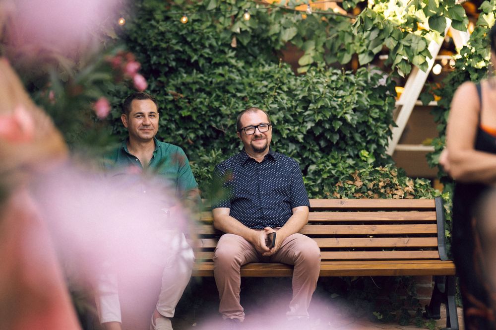 Two men sitting on a wooden bench in a lush garden setting, smiling and relaxed at an outdoor gathering.