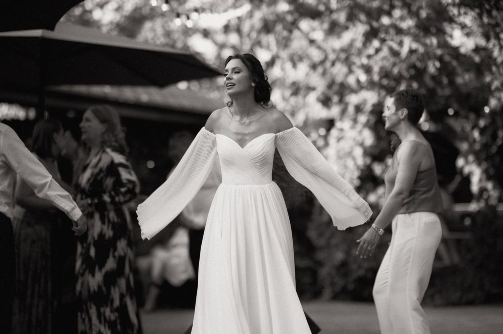 Bride in flowing off-shoulder white gown dancing joyfully outdoors in black and white wedding photo.