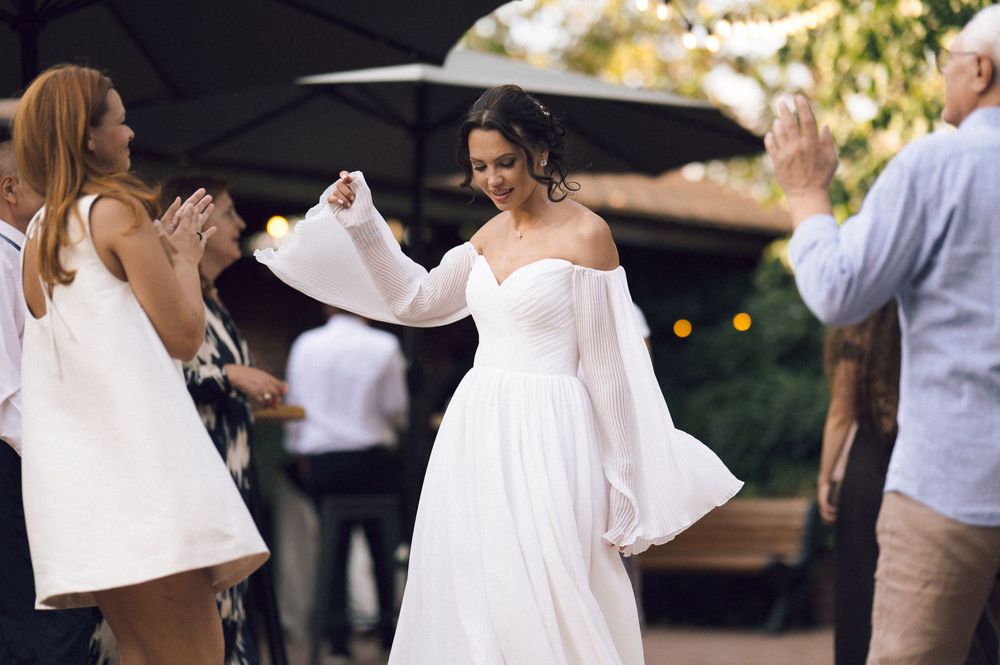 Bride in elegant off-shoulder white gown with flowing sleeves dancing joyfully at outdoor wedding reception.