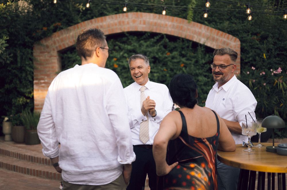 Group of people socializing outdoors at an evening garden party under string lights near a stone archway.