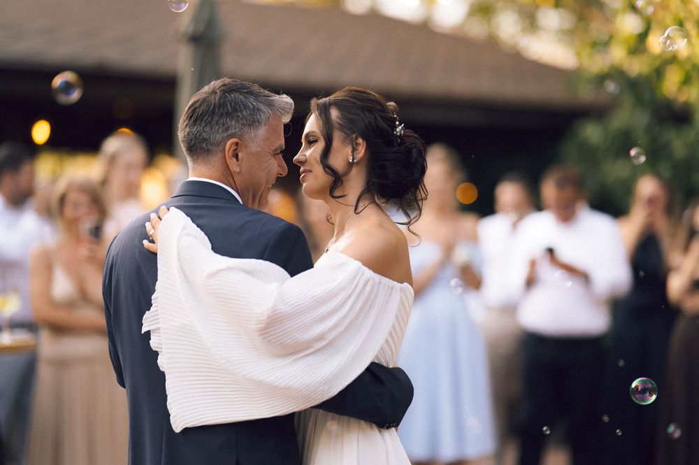 Bride and older man share emotional first dance at outdoor wedding reception with bubbles floating around them.