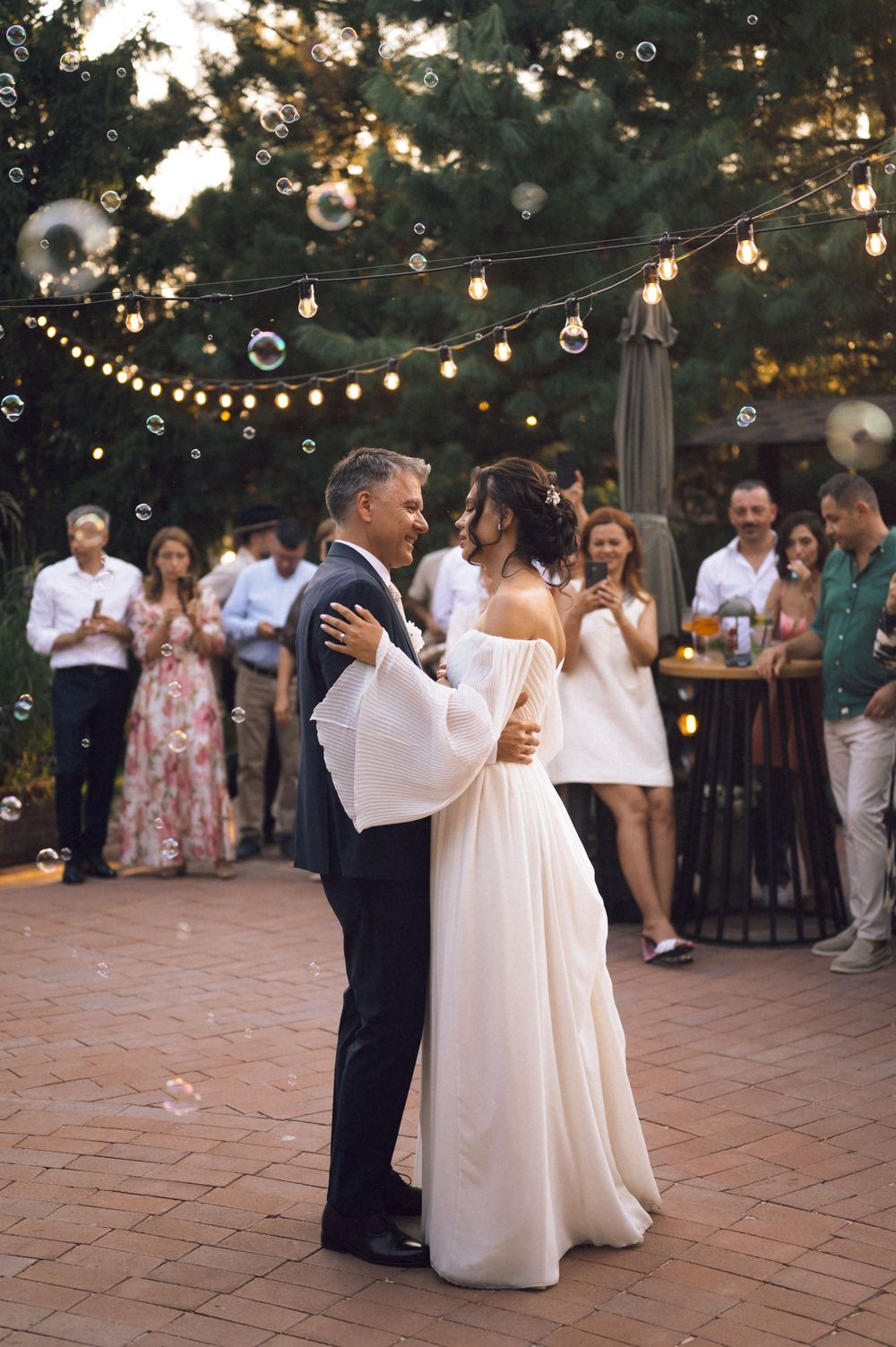 Bride and groom share first dance outdoors under string lights and bubbles with wedding guests watching in background.