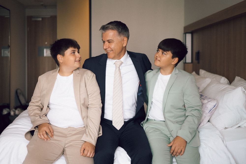 A man in a suit sits on a bed smiling with two boys in blazers in a bright hotel room.
