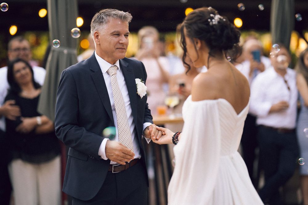 Elegant wedding reception scene with groom in navy suit dancing with bride in white off-shoulder gown amid bubbles.