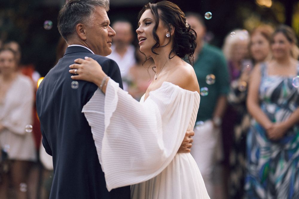 Bride in off-shoulder white gown shares a dance with a man in a suit at an outdoor wedding reception.