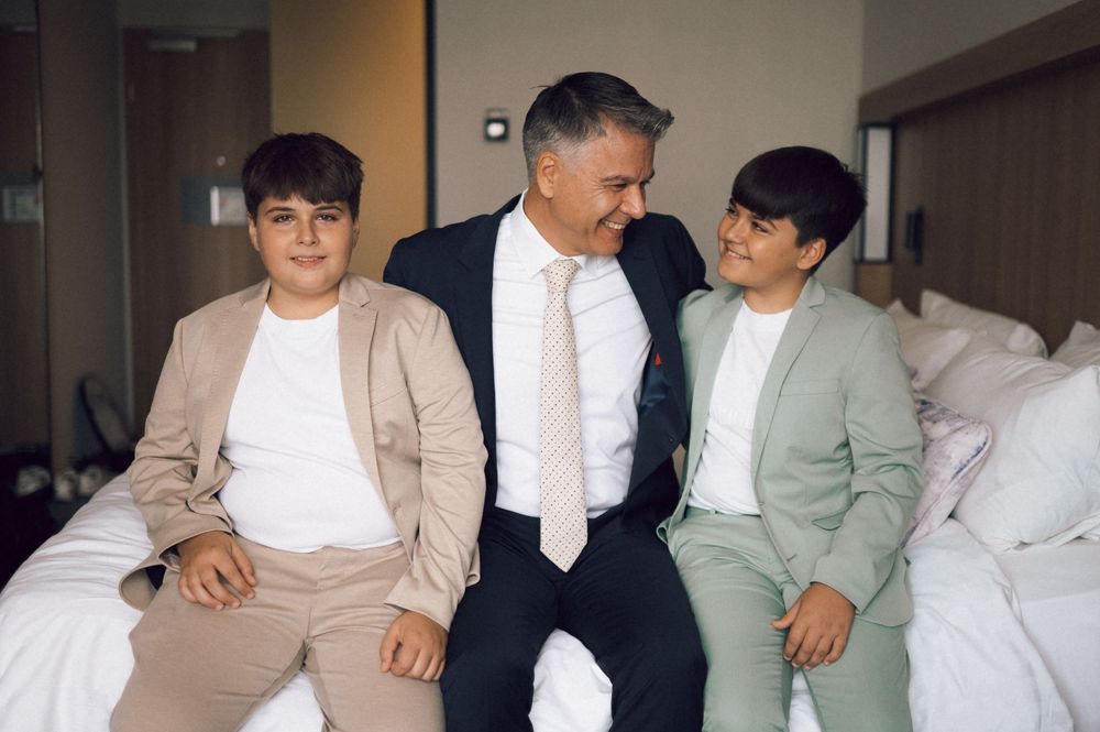 Man in suit sits smiling with two boys in suits on a bed in a hotel room.