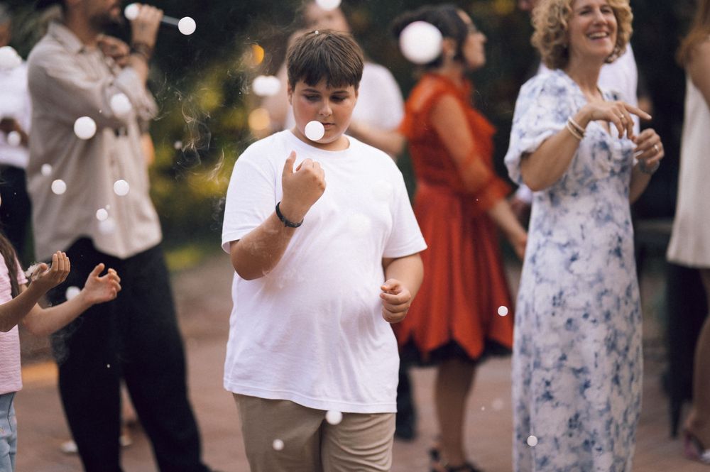 A boy in a white t-shirt blows bubbles at an outdoor evening celebration with guests in the background.