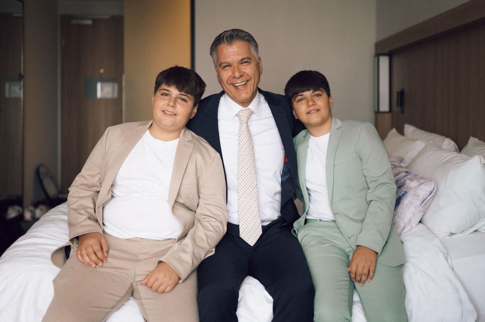 A man in a suit poses with two boys in matching suits on a hotel bed, smiling for a formal family photo.