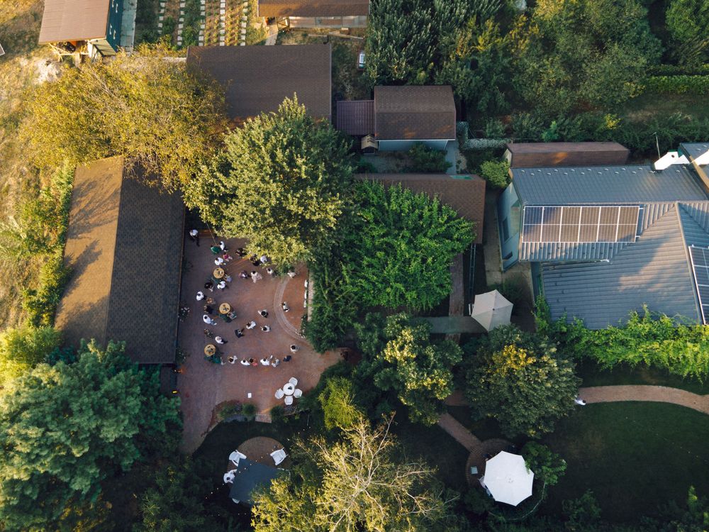 Aerial view of a lush garden venue with outdoor seating, umbrellas, and solar panels surrounded by green trees.