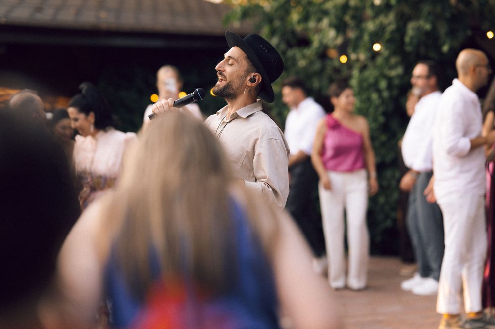 Man in black hat singing at an outdoor party with guests in white attire gathered around him.