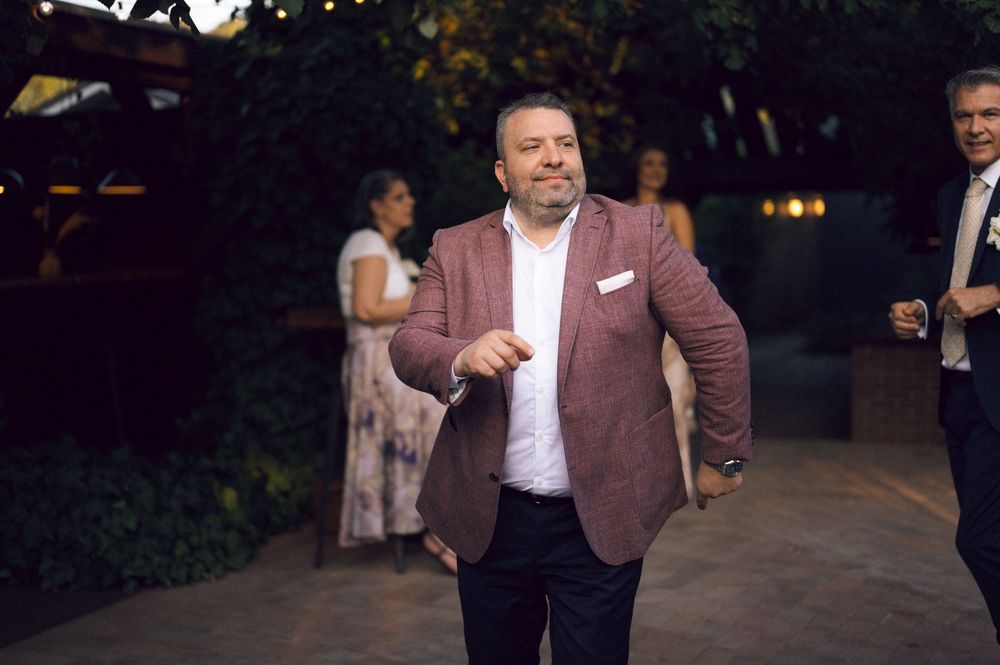 Man in pink blazer pointing at camera at an outdoor evening event with guests in background.
