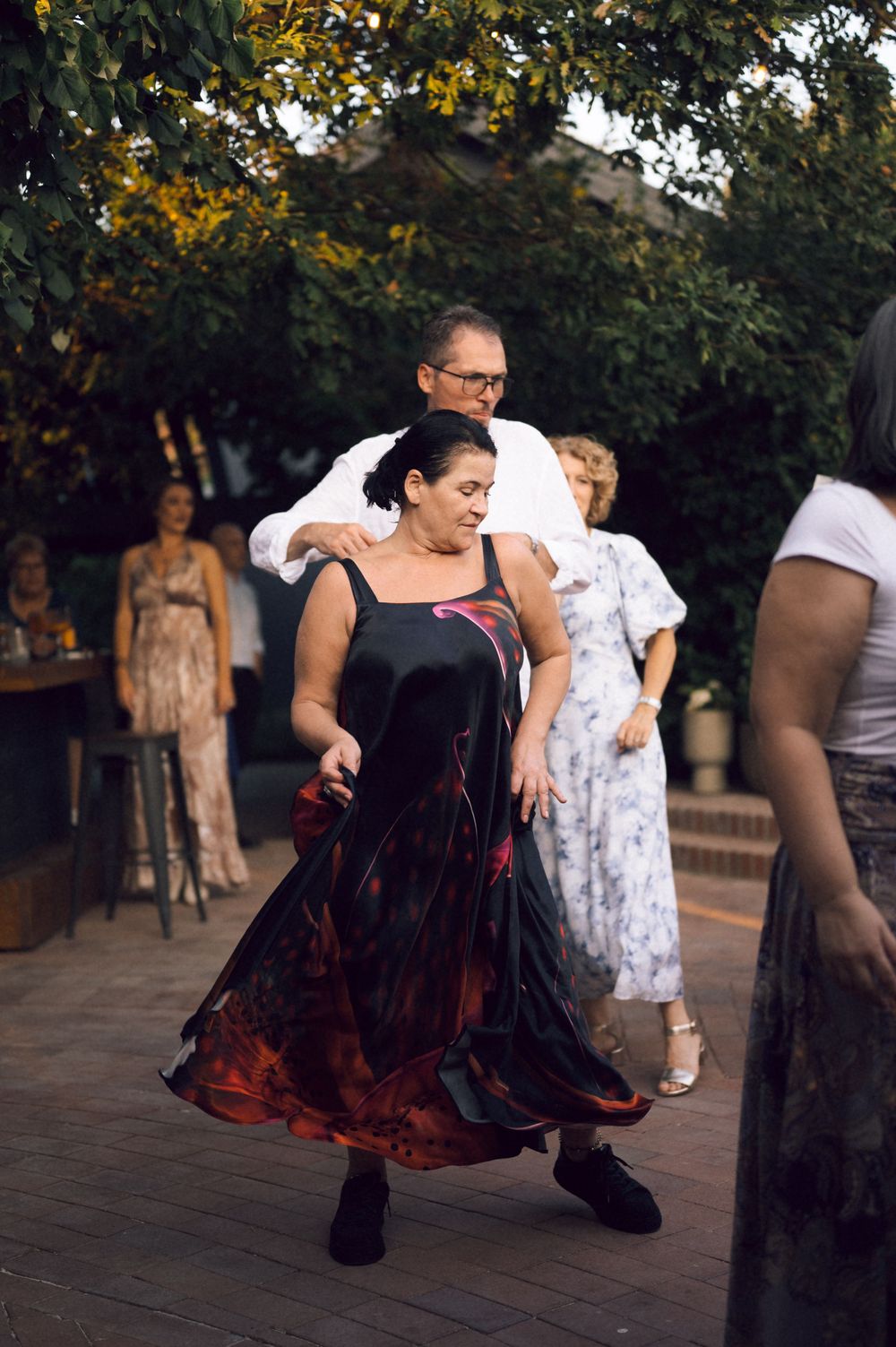 Woman in flowing black and red dress dancing outdoors at a party with guests watching in background.