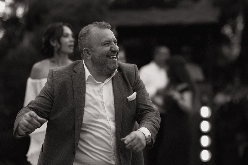 Black and white photo of a smiling man in a suit jacket dancing at an outdoor wedding reception.