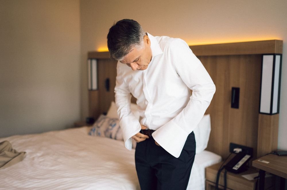 Middle-aged man in white shirt bending over holding stomach in pain while standing beside a bed in a hotel room.