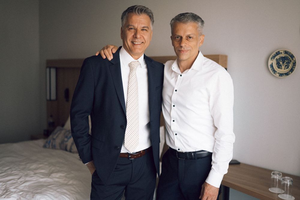 Two well-dressed men smiling and posing together in a neatly decorated bedroom setting.