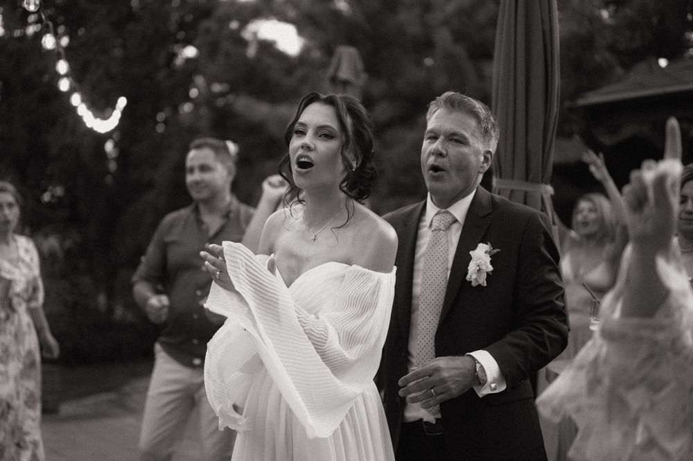 Bride and father share emotional moment singing together at outdoor wedding reception in black and white photo.