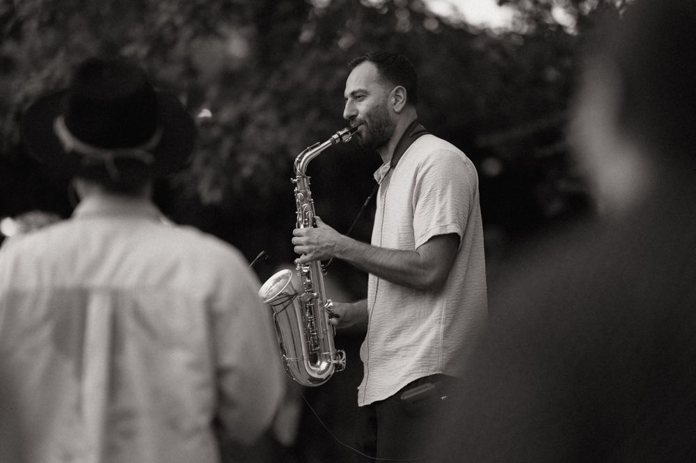 A man plays saxophone outdoors in black and white photo, with blurred crowd in background.