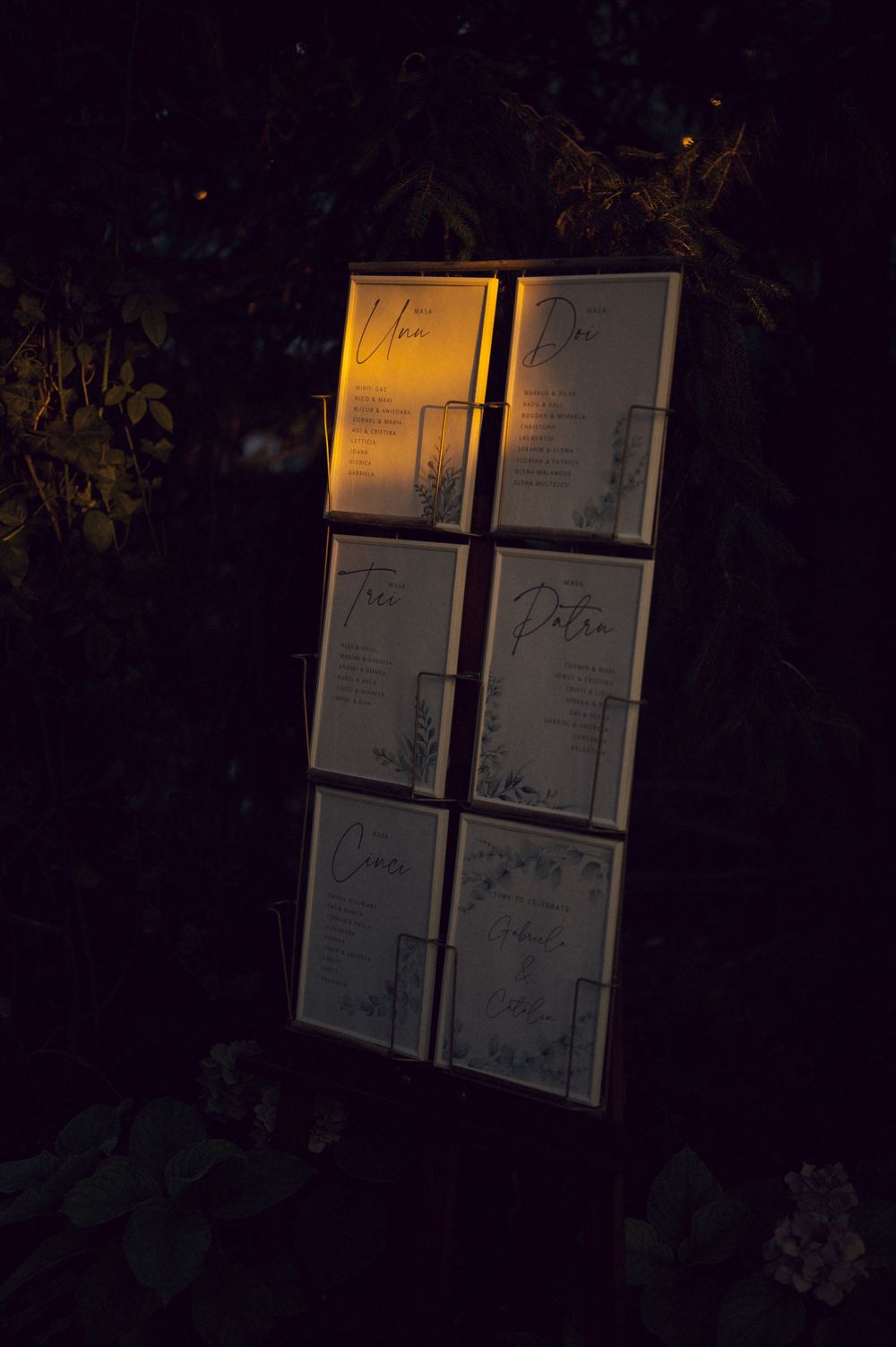Illuminated wedding seating chart display with six framed signs arranged on a dark outdoor stand at night.