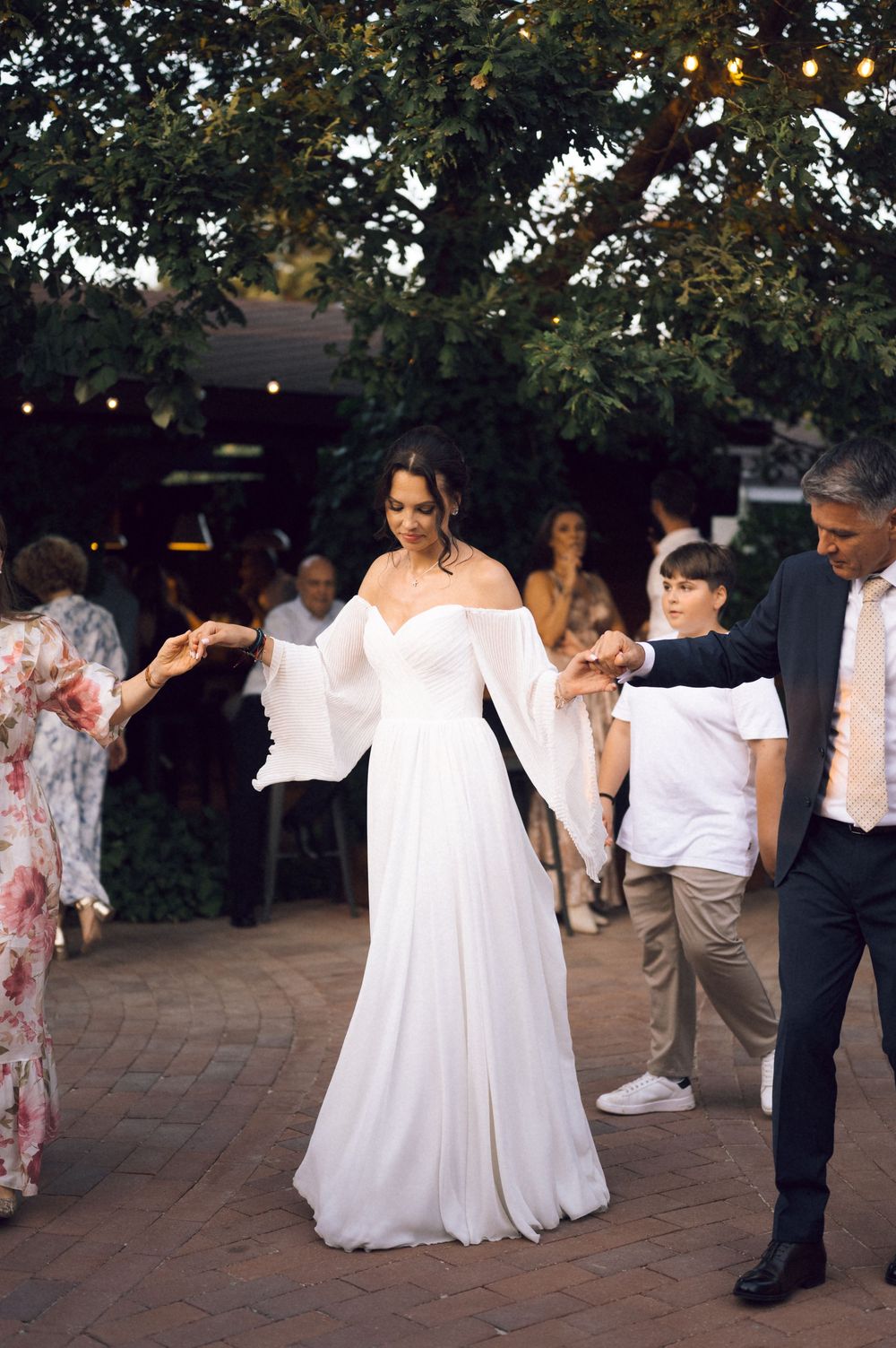 Bride in white off-shoulder gown with bell sleeves dancing outdoors at evening wedding reception.