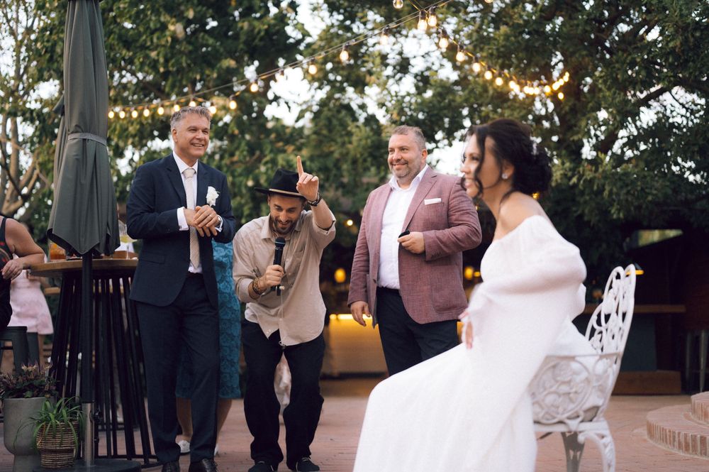 Wedding guests laughing as a performer entertains the bride at an outdoor reception with string lights.