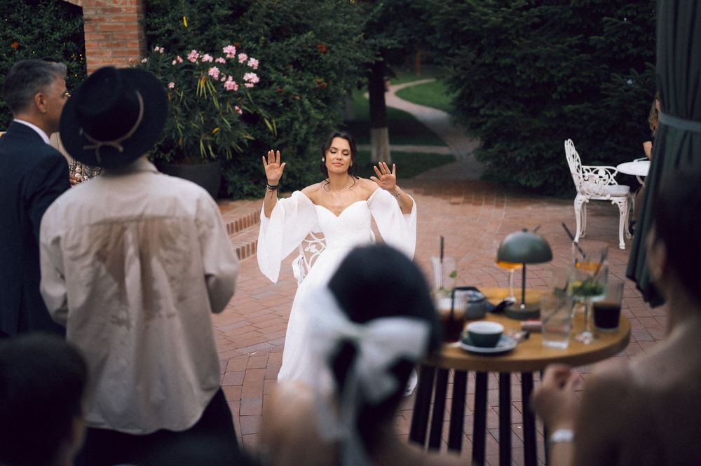 Bride in white off-shoulder gown gestures to outdoor wedding guests on brick patio surrounded by lush greenery.
