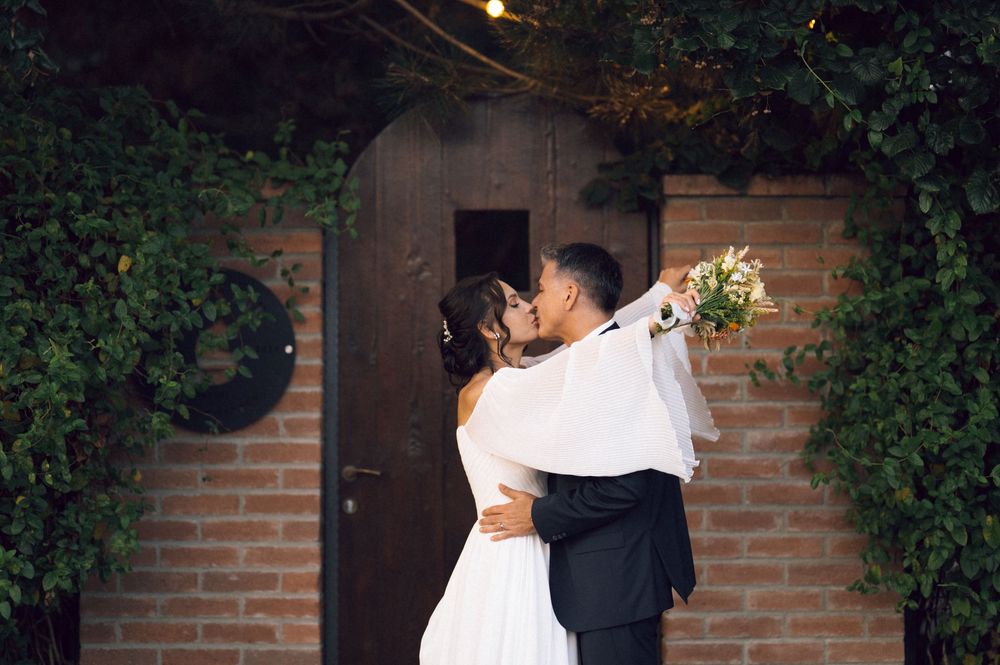 Newlywed couple sharing a romantic kiss in front of a brick wall and wooden door surrounded by lush green ivy.