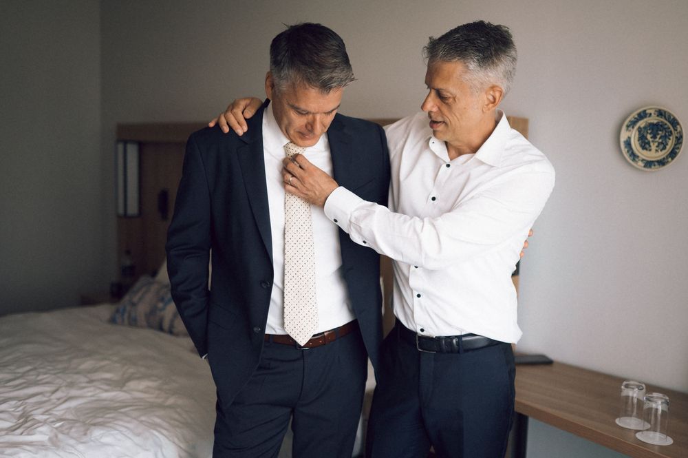 Two men in formal attire stand in a bedroom, one adjusting the other's suit jacket and tie.