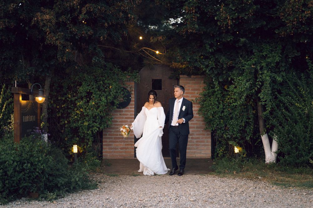 Bride and groom walking hand in hand at night under lush green archway with warm glowing lights at elegant outdoor venue.