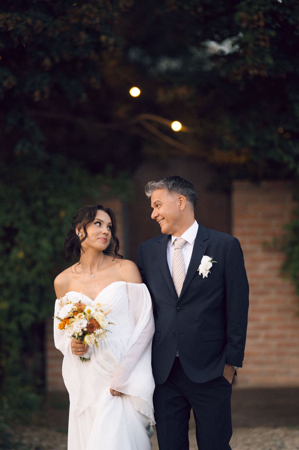 Bride in white off-shoulder gown holding bouquet smiles at groom in dark suit with boutonniere outdoors at evening wedding.