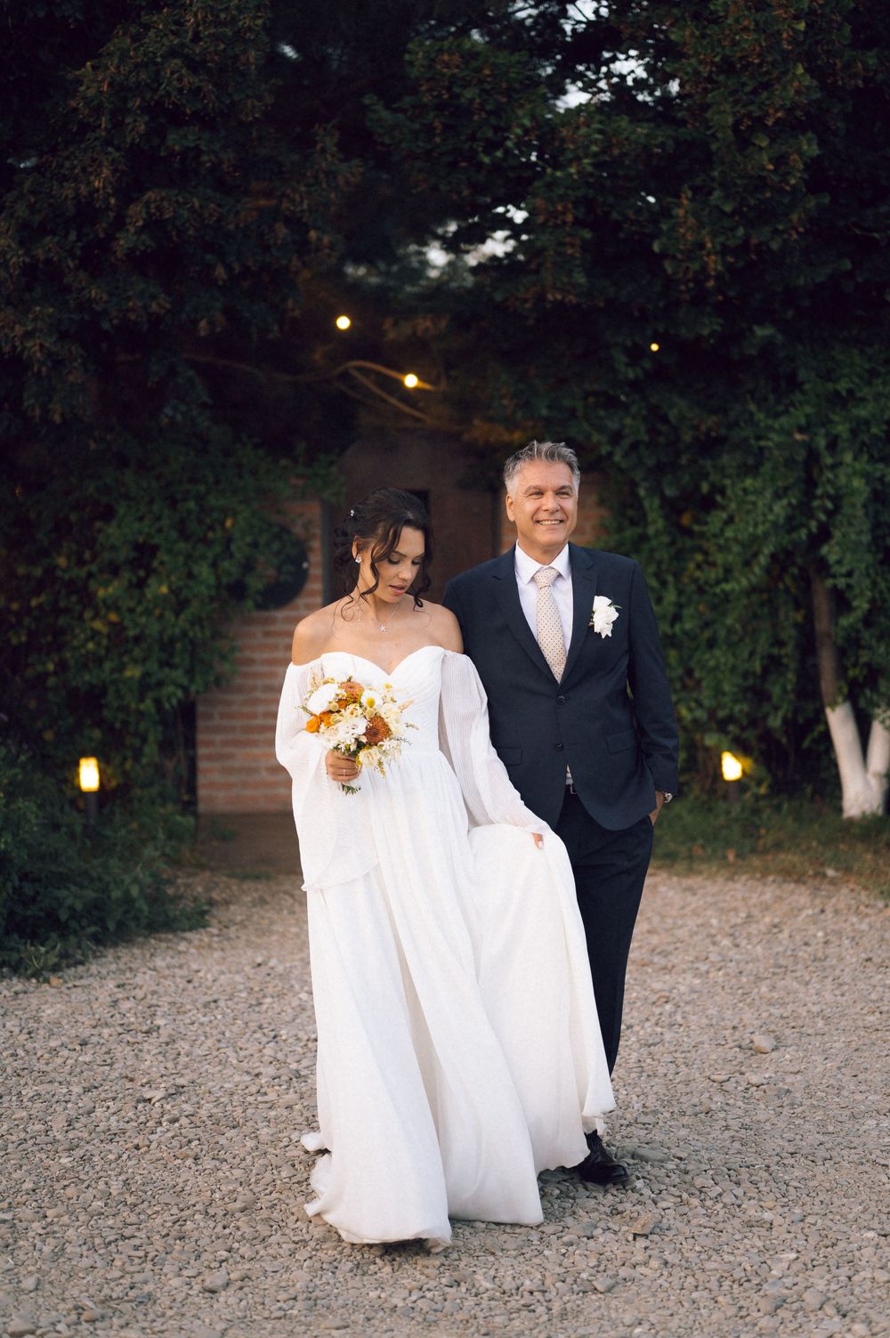Bride in white off-shoulder gown holding bouquet walks with suited man on gravel path surrounded by lush green trees.