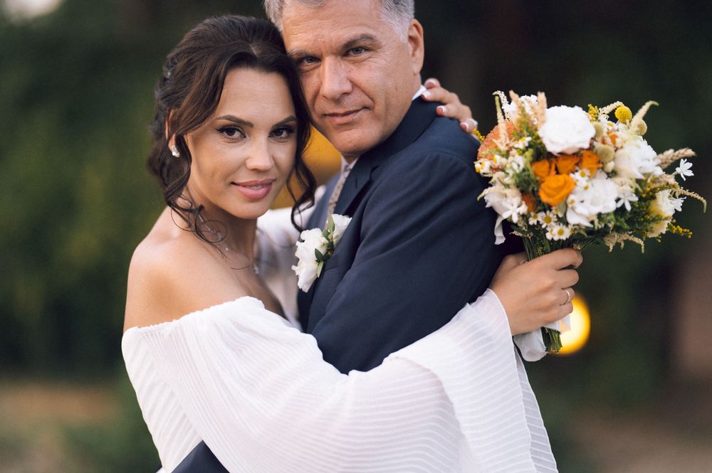 Elegant wedding couple embracing outdoors, bride in white off-shoulder dress holding orange and white floral bouquet.