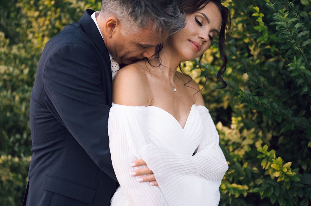Bride and groom embrace tenderly outdoors, she wears an off-shoulder white gown, he wears a dark suit.