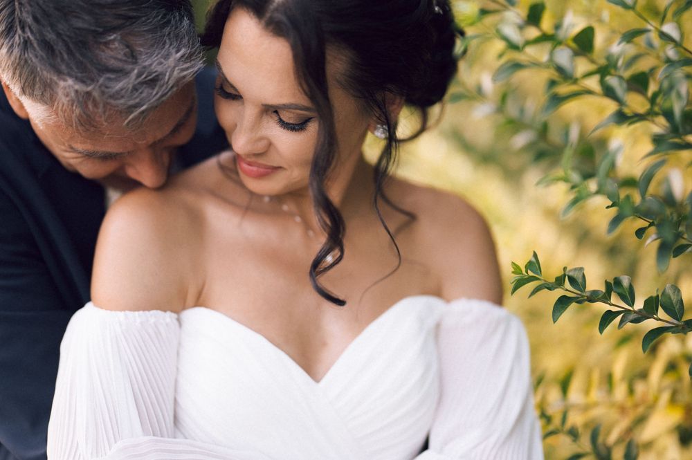 Bride in off-shoulder white gown with groom nuzzling her neck outdoors near lush green foliage.