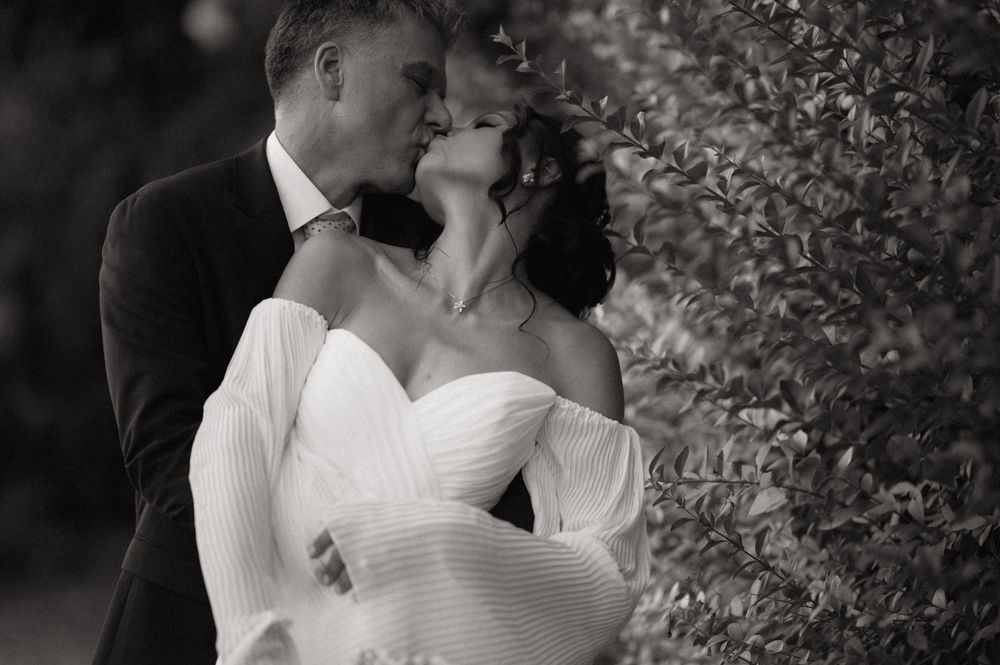 Black and white photo of a couple sharing a romantic kiss on their wedding day beside lush green hedges.