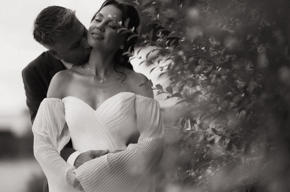 Black and white wedding photo of a couple embracing near lush greenery, bride in off-shoulder gown, groom kissing her cheek.