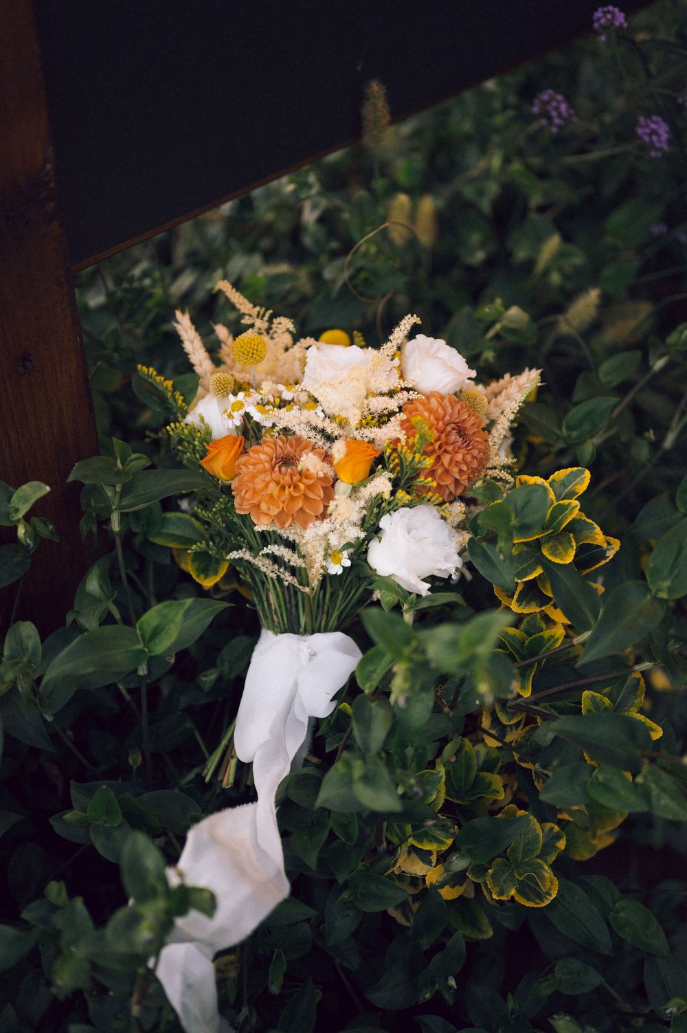 Rustic wedding bouquet with orange dahlias, white peonies, and wildflowers tied with white ribbon, resting on green foliage.