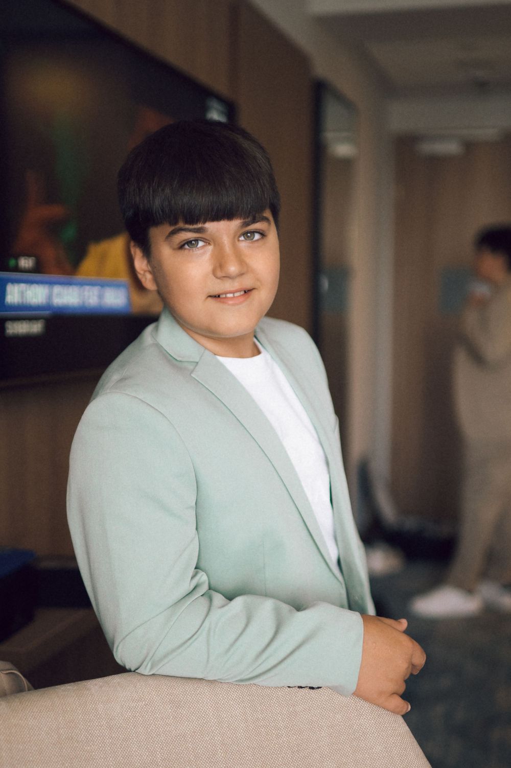 A young boy in a mint green blazer over a white shirt smiles warmly while posing in a modern indoor setting.