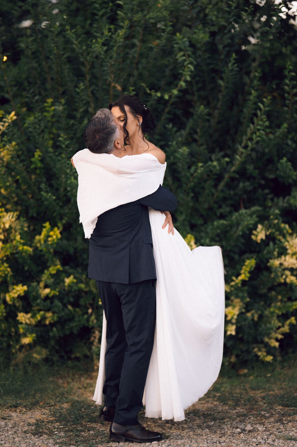 Groom in black suit lifts bride in white off-shoulder gown, sharing a kiss outdoors near lush green hedges.