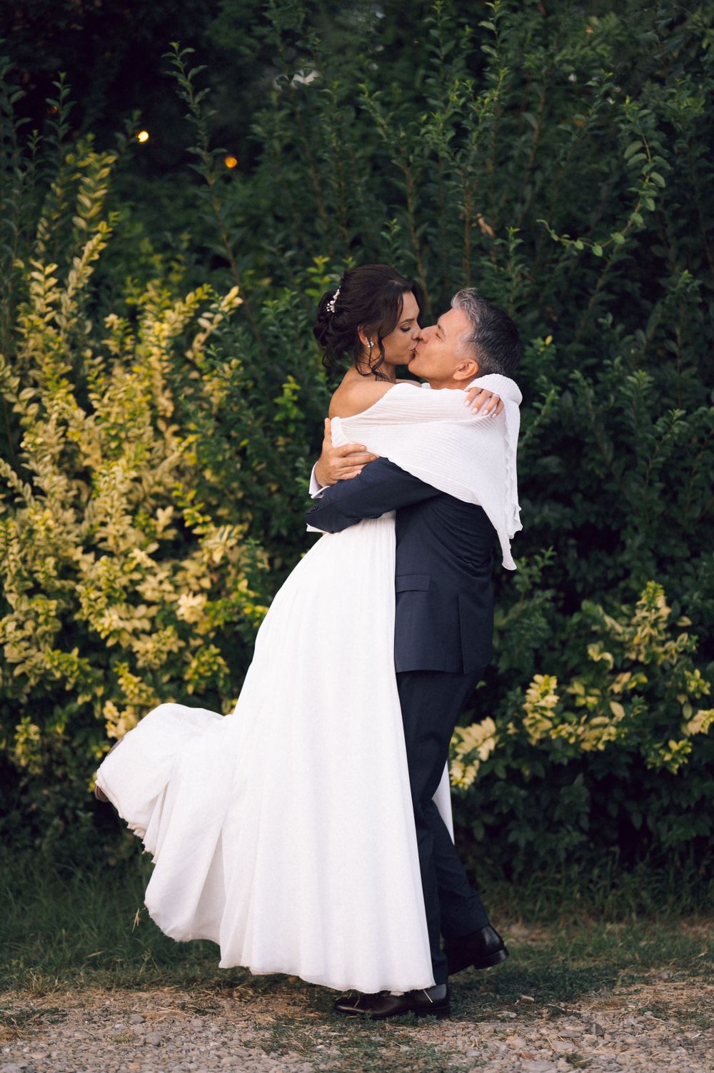 A bride and groom share a romantic kiss outdoors, with the groom lifting his bride in a white gown near lush green bushes.
