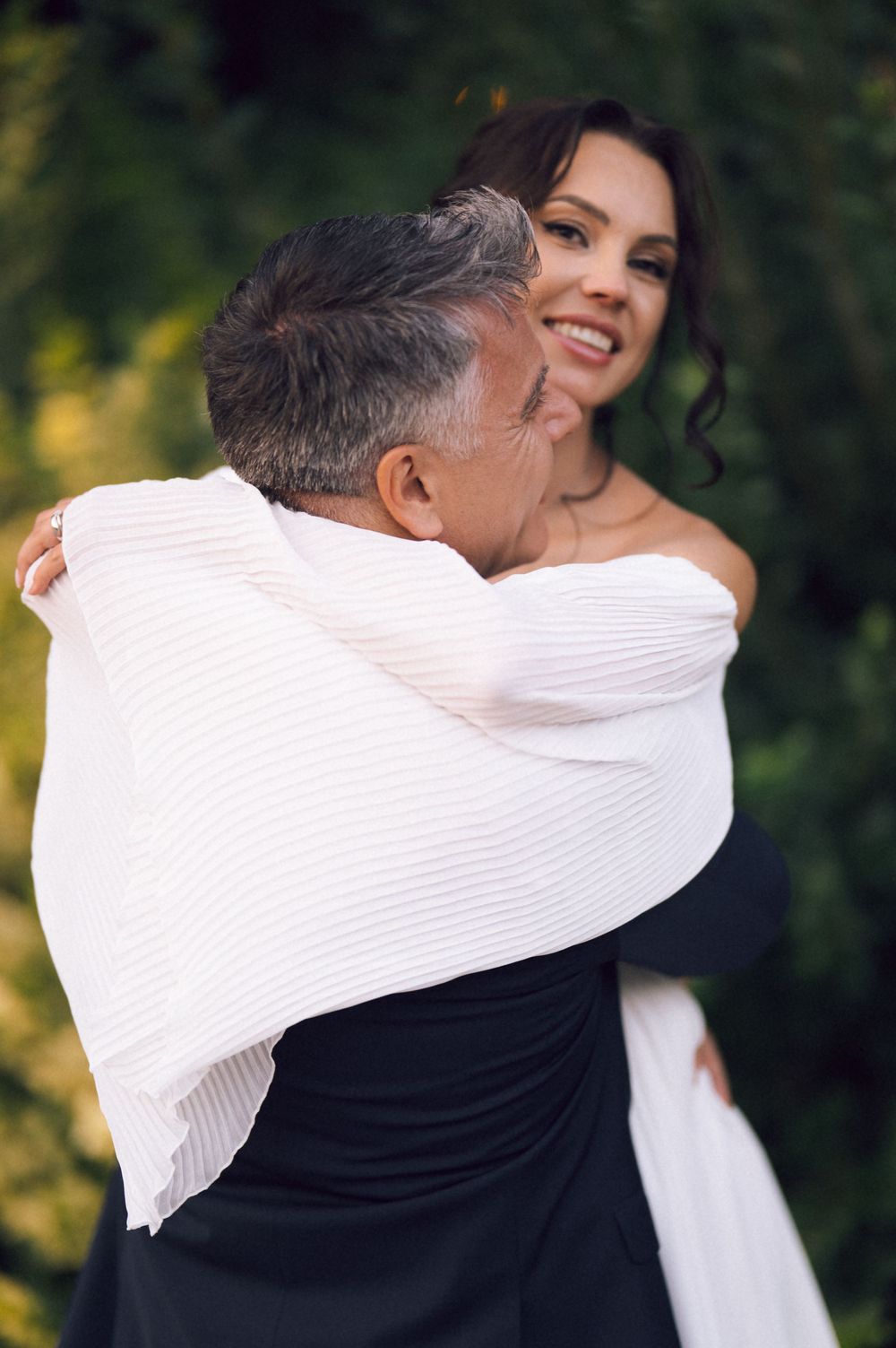 A smiling bride in a white dress shares a warm embrace with an older guest outdoors.